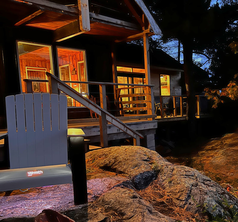 Wide view angle of a Modern Muskoka Chair with Slate legs next perched on rocky ledge with a lit cabin behind